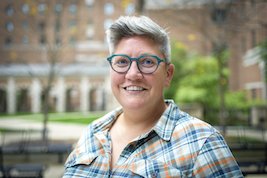 Headshot of Libby-a woman with short dark brown hair, glasses, wearing a plaid top and standing outside